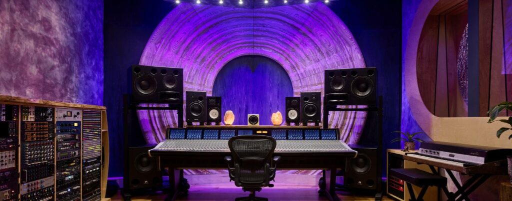 Console de mixage, moniteurs studio et racks d'effets devant un panneau acoustique circulaire éclairé en violet.